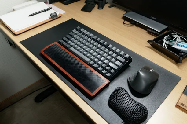 黒いデスクマットの上にキーボードと木製パームレスト、マウスを置いたデスク